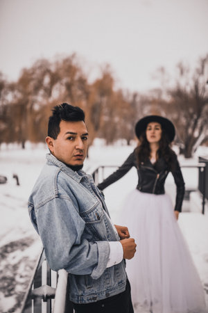 Mexican Hispanic man in oversized denim jacket and woman in hat leather biker jacket kissing hugging laughing walking.groom bride wedding love couple in winter snow.fashionable stylish party weddingの写真素材