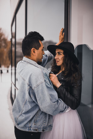 Mexican Hispanic man and woman in hat are kissing hugging laughing walking. groom bride wedding love couple in winter in snow on background of shop windows.fashionable stylish party wedding wearの写真素材