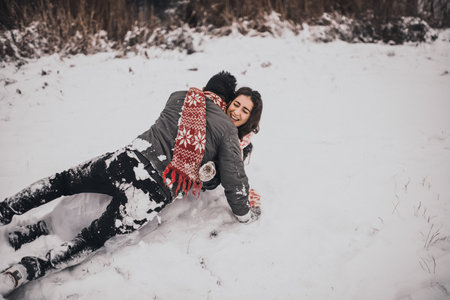 young man and woman running lying in the snow laughing fooling around having fun playing snowballs.bride and groom in love couple in knitted scarf and mittens in winter and snowflakes. mexican peopleの写真素材