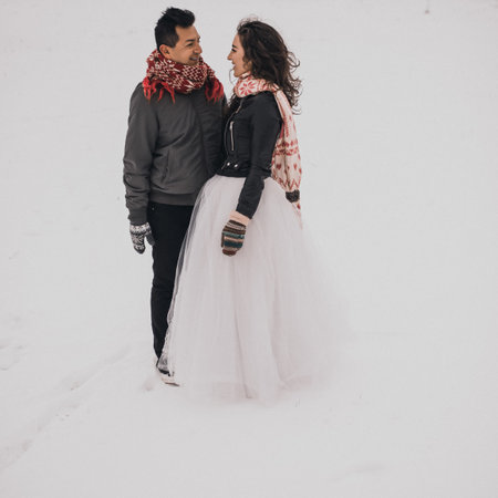 young man and woman stand kissing and hugging. bride and groom in love couple in knitted scarf and mittens in winter with snow and snowflakes. wedding dress. mexican peopleの写真素材