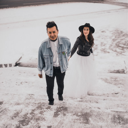 Mexican Hispanic man in oversized denim jacket and woman in hat leather biker jacket kissing hugging laughing walking.groom bride wedding love couple in winter snow.fashionable stylish party weddingの写真素材