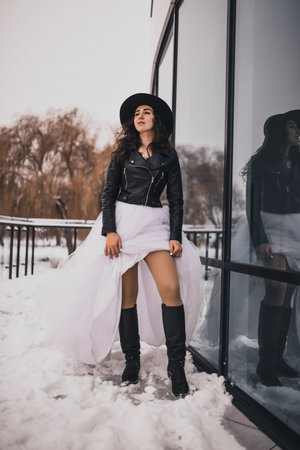 brunette bride woman in wedding white dress, black leather biker jacket and black hat in high boots in winter on the snow. stylish fashion clothing elegant 2021の写真素材