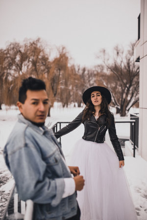 Mexican Hispanic man in oversized denim jacket and woman in hat leather biker jacket kissing hugging laughing walking.groom bride wedding love couple in winter snow.fashionable stylish party weddingの写真素材