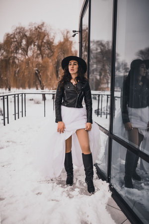 brunette bride woman in wedding white dress, black leather biker jacket and black hat in high boots in winter on the snow. stylish fashion clothing elegant 2021の写真素材