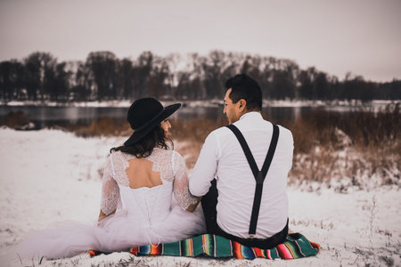 Mexican bride and groom in wedding clothes and suspenders sit on the snow in winter. woman in dress...