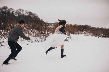 young man and woman running lying in the snow laughing fooling around having fun playing snowballs.bride and groom in love couple in knitted scarf and mittens in winter and snowflakes. mexican peopleの写真素材