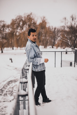 Hispanic brunette man with beard in denim jacket in winter in the middle of snow. Men's Street Fashion Casual Style 2021の写真素材