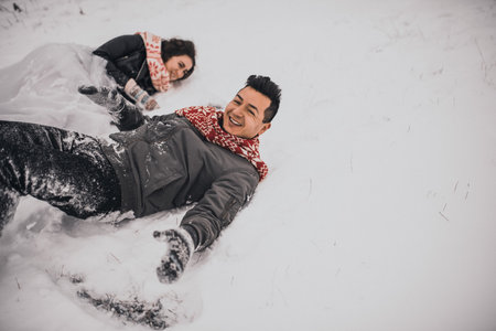 young man and woman running lying in the snow laughing fooling around having fun playing snowballs.bride and groom in love couple in knitted scarf and mittens in winter and snowflakes. mexican peopleの写真素材