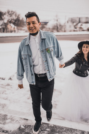 Mexican Hispanic man in oversized denim jacket and woman in hat leather biker jacket kissing hugging laughing walking.groom bride wedding love couple in winter snow.fashionable stylish party weddingの写真素材