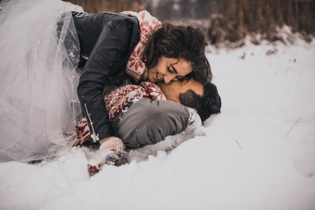 young man and woman stand kissing and hugging. bride and groom in love couple in knitted scarf and mittens in winter with snow and snowflakes. wedding dress. mexican peopleの写真素材