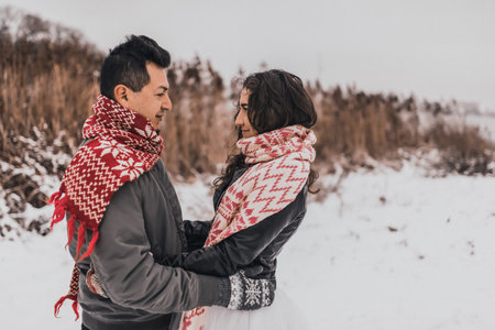 young man and woman stand kissing and hugging. bride and groom in love couple in knitted scarf and mittens in winter with snow and snowflakes. wedding dress. mexican peopleの写真素材