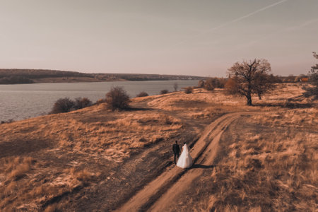 The bride and groom in wedding dresses walk along the autumn red field near the river. Newlyweds in nature. white wedding dress with veil and black suit for men. Top viewの写真素材