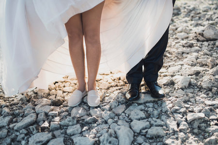 Legs of the bride and groom standing on gray small stones. white light wedding dress and black trousers for men. Men's classic shoes and sports pink shoes on the brideの写真素材