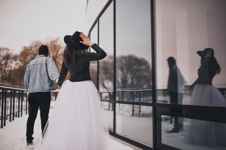 A man in a light blue denim jacket is leading his black-haired curly-haired bride in a white dress with a black leather jacket and a wide-brimmed black hat. Snow lies near the mirror showcases.の写真素材
