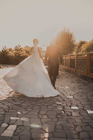 The wedding couple runs away into the sunset.The bride in a white long wedding dress is walking by the hand with the groom in a business classic suit. White sky dark gray brick floor on the backgroundの写真素材