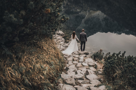 a couple of light-skinned newlyweds In wedding clothes. a large transparent lake and gray stones and trees. long-haired brunette bride in a white long dress and a slim man groomの写真素材