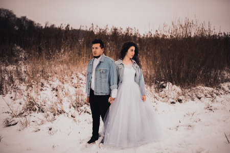 Mexican Hispanic man in denim jacket oversized and woman are kissing hugging laughing. groom and bride wedding love couple in winter in snow. fashionable stylish party wedding wear 2021の写真素材