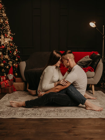 in love couple man and woman sitting on floor cross-legged embrace hugging and kiss Christmas tree. decorated house for New Year. Christmas morning. apartment interior. Valentine's day celebrationの写真素材