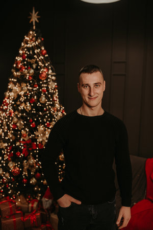 handsome young man in black clothes near a Christmas tree in garlands. decorated house for New Year. Christmas morning. apartment interior. Valentine's day celebrationの写真素材