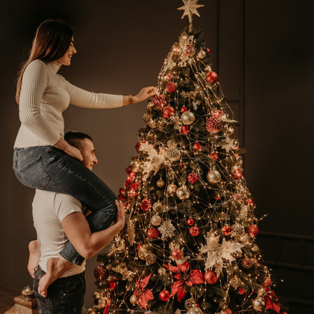 young woman sits on back shoulders of man and decorates a Christmas tree, hangs ornaments. decorated house for New Year. Christmas morning. apartment interior. Valentine day celebrationの写真素材