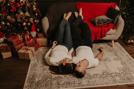 young couple man and woman lie on floor near couch with legs feet up hugging kissing in the bedroom near christmas tree. decorated house for New Year. Valentine's day celebrationの写真素材