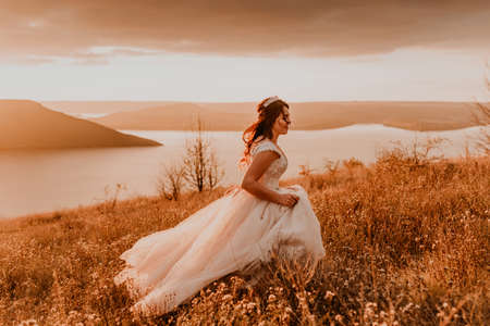 beautiful woman brunette bride in white dress and suit are walking on tall grass in field in summerの写真素材