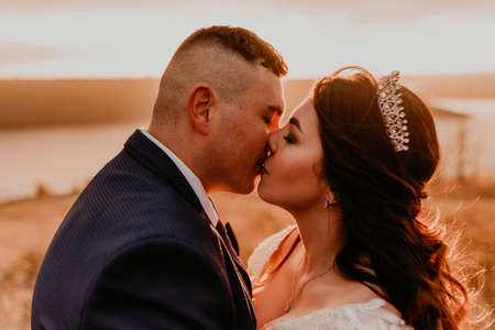 A loving couple wedding newlyweds in white dress and suit walk hug kissing whirl on tall grass in summer field on mountain above the river.style bride accessories on head crown makeup hairstyleの写真素材
