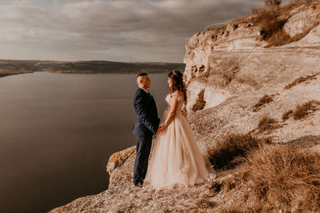 loving couple wedding newlyweds in white dress and suit walk in summer on mountain above river. sunset and sunrise. man and woman on rocks above cliffの写真素材
