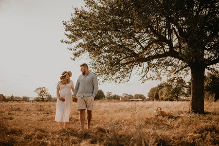 Happy family pregnant caucasian blonde woman in hat in white cotton dress walks with husband in meadow in summer. man in light natural clothes and shorts holds hand wife. trees in sunbeamsの写真素材