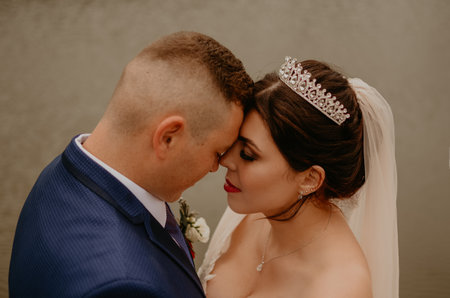 blonde European Caucasian young man groom in blue suit and black-haired woman bride in white wedding dress with long veil and tiara on head. Newlyweds hold hands smile kiss and look at each other near riverの写真素材