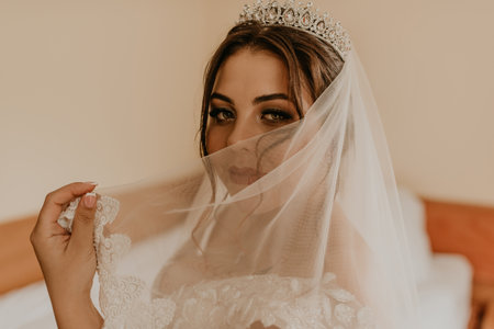 European Caucasian young black-haired woman bride in white wedding dress with long veil and tiara on head. girl bedroom in beige colors morning gatherings of preparing bride for holiday party weddingの写真素材