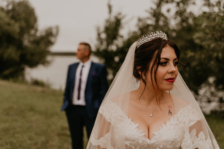 European Caucasian young man groom in blue suit and black-haired woman bride in white wedding dress with long veil and tiara on head. newlyweds look in different directions. man stands behind womanの写真素材