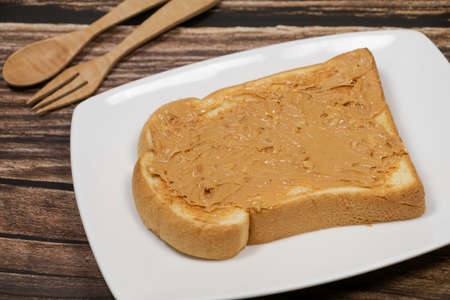 Sliced bread spreading with peanut butter with spoon and fork on white plate. on wooden boardの写真素材