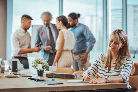 Smiling businesswoman writing in a notebook in the office with colleagues in the backgroundの写真素材
