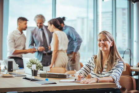 Attractive businesswoman sitting at her desk in the office with colleagues in the backgroundの写真素材