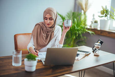 Portrait of beautiful muslim businesswoman in hijab working with laptop and drinking coffee at officeの写真素材