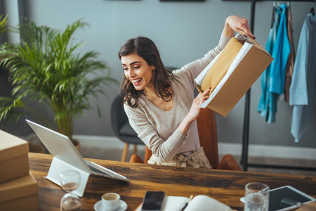 Cheerful young woman unpacking parcel box and smiling while sitting in office