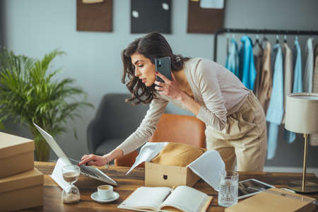 Young woman in casual clothes is using a laptop and talking on the phone while working in the office.の写真素材