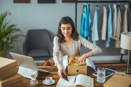 Beautiful young woman packing a parcel to her home office, she is using a laptop and smilingの写真素材