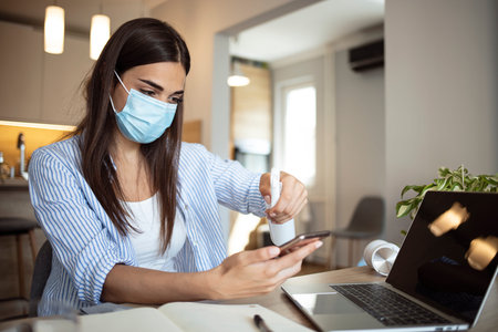 Young woman in protective mask using smartphone and sanitizer at home