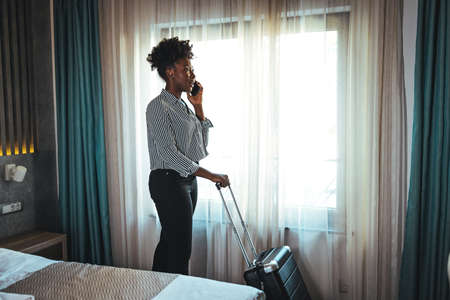 Young african american businesswoman talking on the phone while standing with suitcase in hotel roomの写真素材