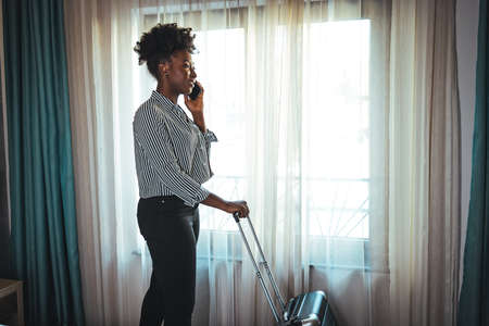 Afro american businesswoman with suitcase talking on the phone in hotel roomの写真素材