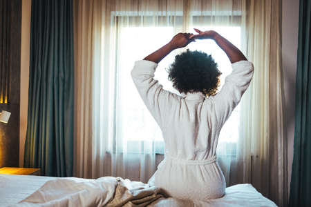 Back view of young african american woman in white bathrobe stretching on bed at home.の写真素材