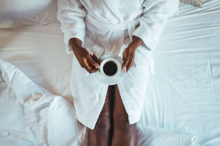 Top view of african american woman in bathrobe holding cup of coffee while sitting on bedの写真素材