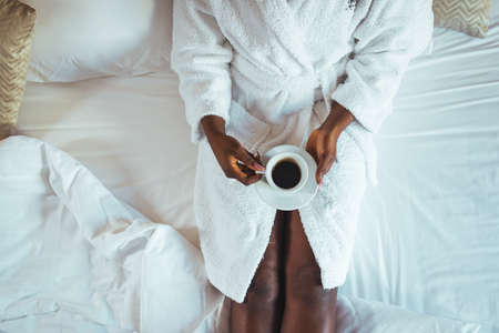 Top view of african american woman in bathrobe holding cup of coffee while sitting on bedの写真素材