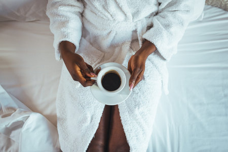 Top view of african american woman in white bathrobe holding cup of coffee on bed