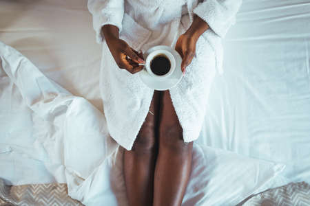 Cropped view of african american woman in bathrobe holding cup of coffee on bedの写真素材