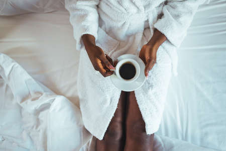 Cup of coffee in hands of african american woman in white bathrobe on bedの写真素材