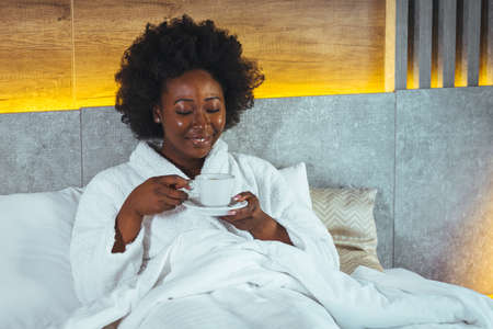 Young woman in bathrobe drinking coffee in hotel room. Attractive Afro american lady relaxing in hotel room.の写真素材