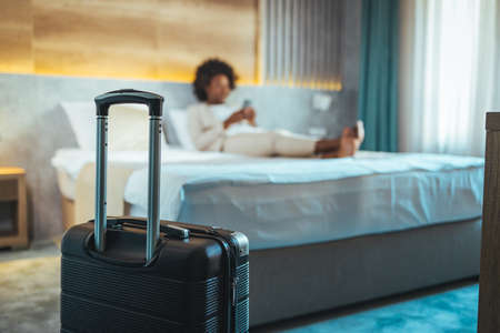 Close-up of black suitcase in hotel room, blurred african american woman lying on bed in backgroundの写真素材